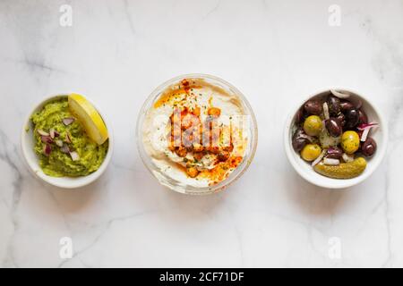 From above tasty healthy hummus green sauce cut onion salted cucumbers decorated with sliced lemon in white bowls on table Stock Photo