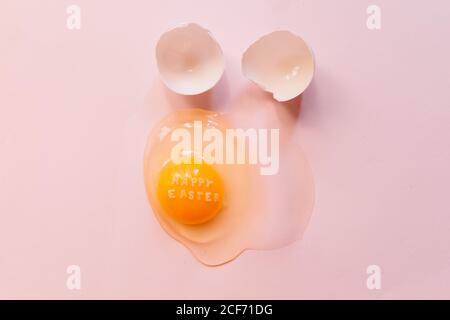 From above raw broken egg with text Happy Easter on yellow yolk and eggshells on pink background Stock Photo