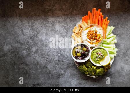 From above appetizing healthy hummus green sauce cut carrot cucumbers and green broccoli salted cucumbers decorated with sliced lemon in white bowls on table Stock Photo