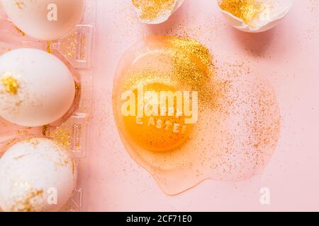 Top view of broken glittered egg with text Happy Easter and raw eggs in plastic box on pink background Stock Photo