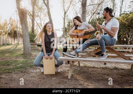 Young man and women with musical instruments sitting near table and laughing on sunny day in park Stock Photo