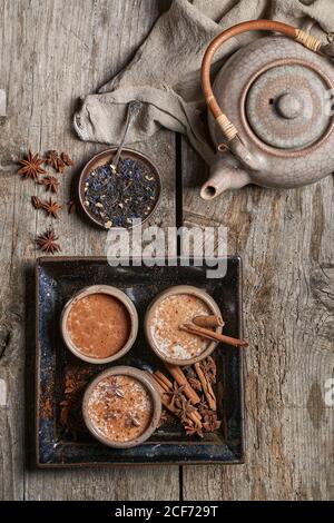 Top view of Masala chai served in ceramic bowls with star anise and cinnamon sticks arranged on wooden table with teapot and piece of cloth Stock Photo