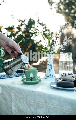 Crop female pouring coffee from metal geyser coffeemaker to green cup while preparing breakfast standing at table with bouquet of wildflowers in vase and fresh donuts with glaze on plate in nature Stock Photo