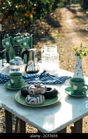 High angle of tasty donuts with icing on plate and cups of hot drink beside tableware set and moka pot in composition with vase with small yellow flowers on table in nature in sunny day Stock Photo