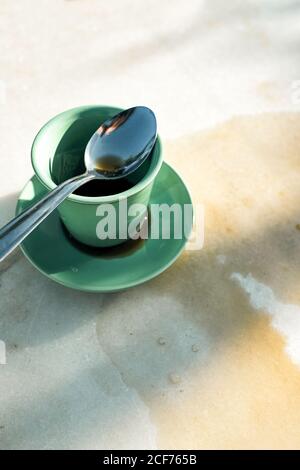 From above green ceramic cup of warm black coffee spilled on table composed with metal teaspoon on saucer on gray table Stock Photo