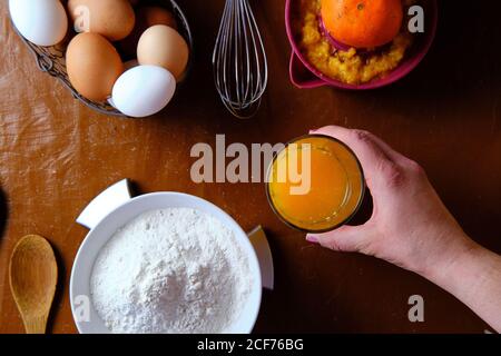 Top view of anonymous mature Woman putting glass of fresh citrus juice on table near bowl with flour and basket of raw eggs during pastry preparation at home Stock Photo