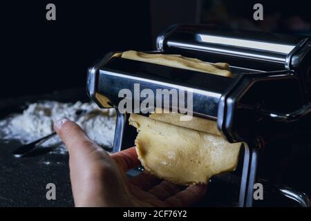 Crop person rolling dough through pasta machine while preparing fresh homemade pasta on table with flour and spoon in background Stock Photo