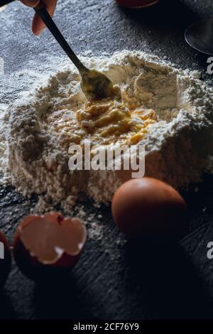 Crop person with fork in hand mixing egg and flour while preparing dough for pasta on black table Stock Photo