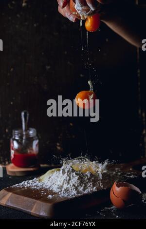 Crop person breaking eggs over flour while preparing dough on table with eggshell and glass pot and egg falling on black background Stock Photo