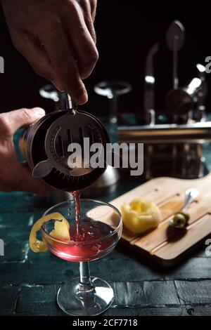 Crop anonymous barman pouring red alcohol cocktail from metal shaker into coupe glass garnished with lemon peel while working at counter in pub Stock Photo