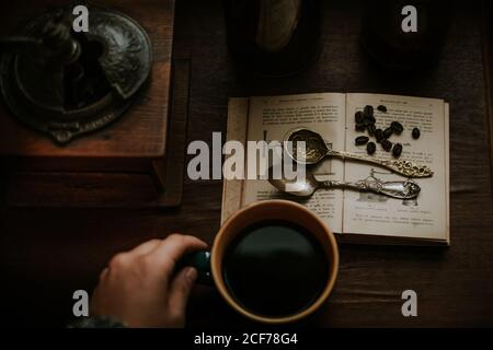 Top view of cropped person hands holding coffee mug near vintage silver spoons and coffee beans placed on open old book near manual coffee grinder Stock Photo