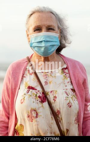 Senior gray haired lady in blue sterile mask and casual clothes looking at camera while standing on blurred background in daylight Stock Photo