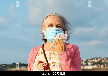 Senior female in sterile mask and casual wear talking phone while standing under blue cloudy sky outdoors during quarantine time Stock Photo