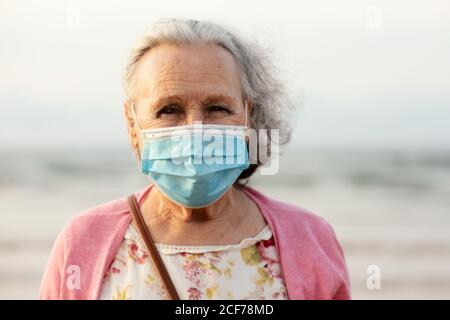 Senior gray haired lady in blue sterile mask and casual clothes looking at camera while standing on blurred background in daylight Stock Photo