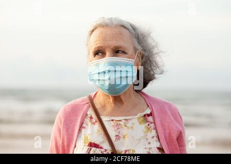 Senior gray haired lady in blue sterile mask and casual clothes looking away while standing on blurred background in daylight Stock Photo