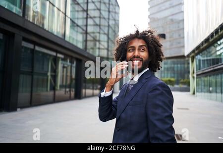 Cheerful African American man in formal suit standing outside and talking on smartphone while looking away Stock Photo