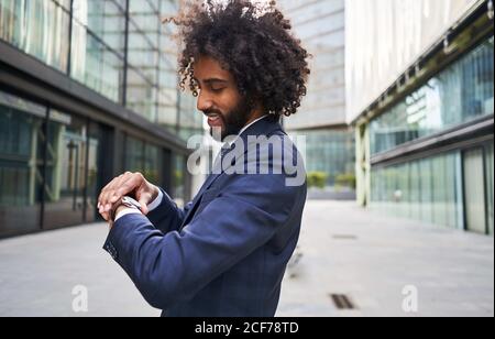 Cheerful black office worker with curly hair standing at street and looking at watch on blurred background Stock Photo