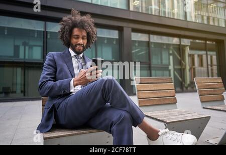 Joyful ethnic man in suit sitting outside and messaging Stock Photo