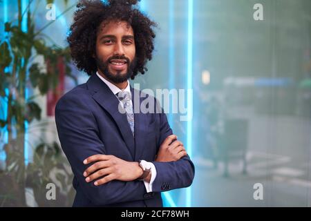 Confident and presentable African American businessman standing with crossed arms near window and looking at camera Stock Photo