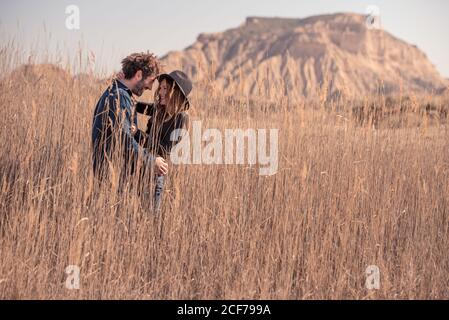 Side view of young man and Woman in casual clothes smiling and embracing each other while standing in tall dry grass near mountain in Bardenas Reales in Navarre, Spain Stock Photo