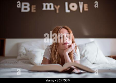 Peaceful female relaxing on bed while lying on stomach and reading interesting book in cozy bedroom with romantic inscription on wall and dreamy atmosphere Stock Photo