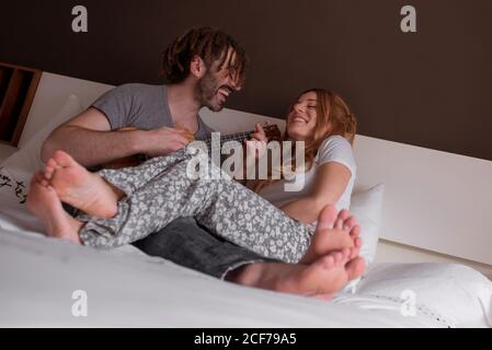 cheerful young man with dreadlocks and Woman with red hair sitting on bed and having fun with closed eyes playing ukulele guitar while spending time together at home at weekend Stock Photo