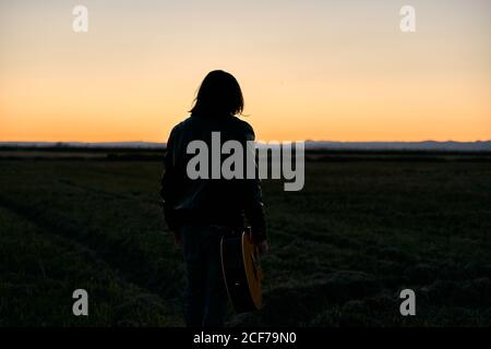 Back view of anonymous dark silhouette of male musician with long hair walking towards camera with guitar in hands at sunset