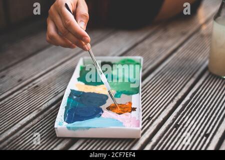 Closeup hand of anonymous artist using brush to mix paint in carton box while spending time on terrace