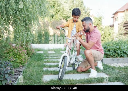 Little boy sitting in bicycle waiting for his father to fix handlebar height outdoors on sunny summer day Stock Photo