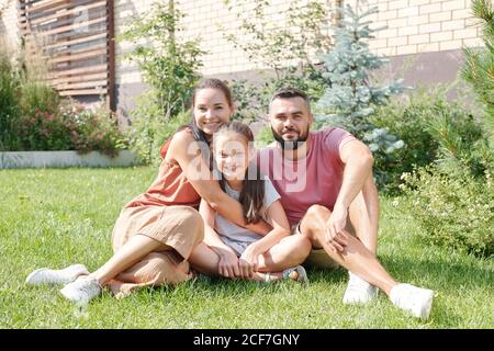 Happy father, mother and daughter spending time together in backyard sitting on lawn smiling at camera Stock Photo