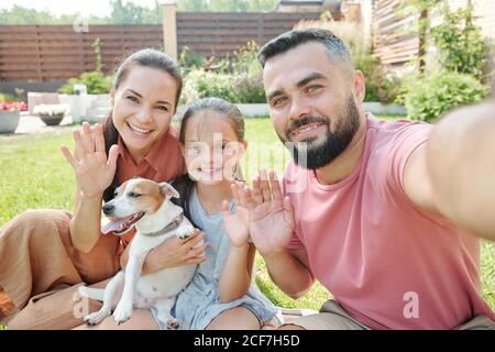 Father taking selfie shot of himself and his family waving hands at camera, girl holding cute dog Stock Photo