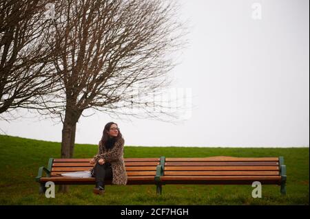 Beautiful female with umbrella looking away while sitting on bench near leafless trees on misty day in quiet park Stock Photo