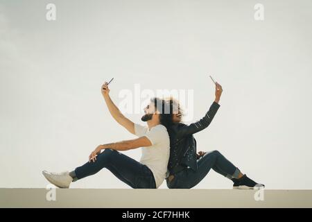 Side view of young multiethnic man and Woman smiling and taking selfies while sitting back to back on building wall against gray sky Stock Photo