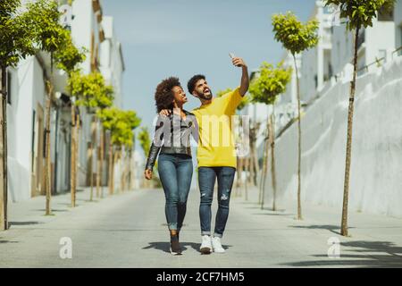 Handsome bearded man and pretty Woman walking while taking selfie on sunny day on city street Stock Photo