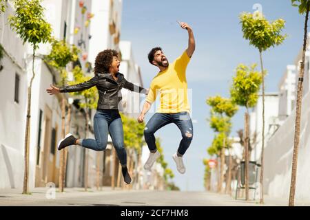 Handsome bearded man and pretty Woman screaming and leaping up while taking selfie on sunny day on city street Stock Photo