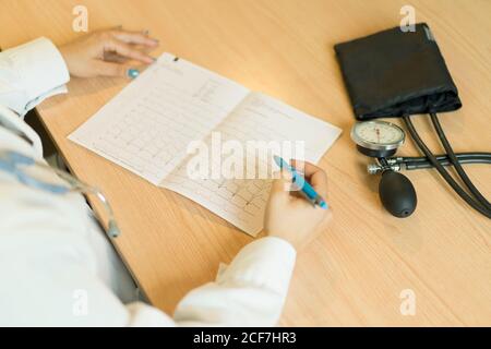anonymous young female doctor in uniform looking at paper and sitting at table with tonometer in room Stock Photo