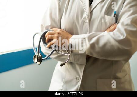 Crop female doctor in uniform with crossed hands holding stethoscope in room Stock Photo