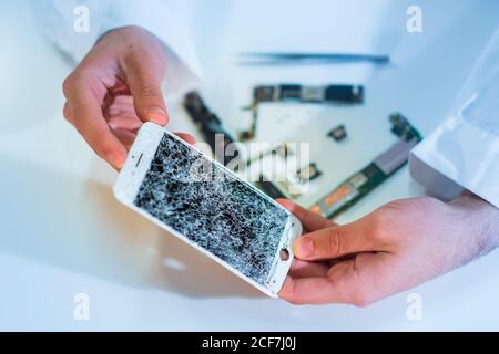 Hands of unrecognizable technician demonstrating cracked screen of modern mobile phone over white table in workshop Stock Photo