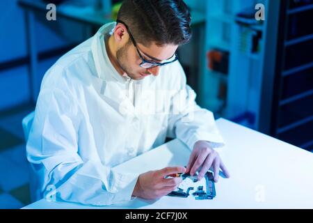 Young technician in glasses and white robe fixing contemporary gadget on white table in workshop Stock Photo