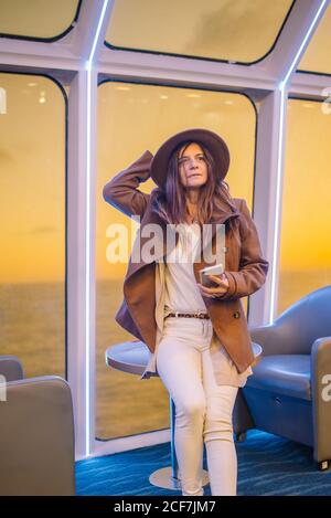 Relaxed stylish young female in trendy outfit sitting on chair using mobile phone on board of luxury cruise ship and enjoying view from window with calm sea water and sunset sky Stock Photo