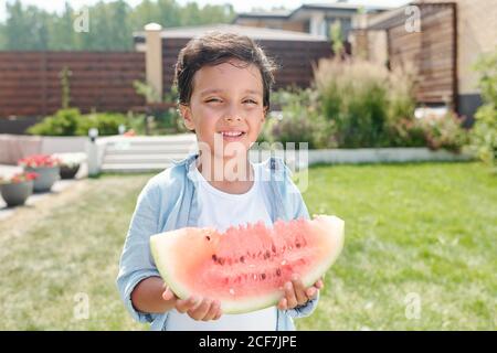 Waist up portrait shot of cheerful little boy standing in backyard holding piece of watermelon smiling at camera Stock Photo