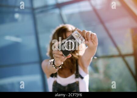 From above of young unrecognizable female holding photo camera in outstretched hands and taking picture while standing against glass wall Stock Photo