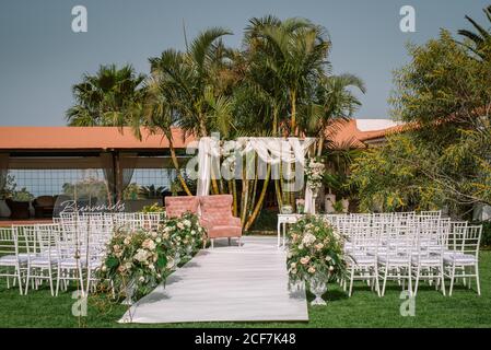 Rows of white chairs walkway among flowers against wedding arch on yard of green tropical resort with palms on sunny day Stock Photo