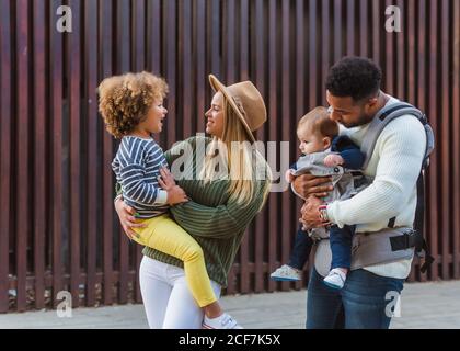 Cheerful stylish young multiracial couple with infant son and little daughter in casual wear walking together along wooden fence on city street Stock Photo