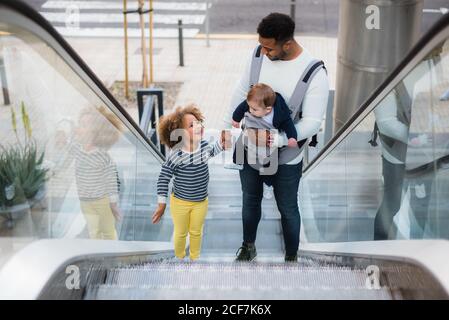 From above of positive young ethnic man carrying infant son and holding hand of cheerful little daughter while standing on escalator in city Stock Photo