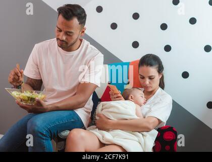 Black haired mother with sleepy baby ob chest with mobile phone in hand sitting on chair with father in casual wear eating salad Stock Photo
