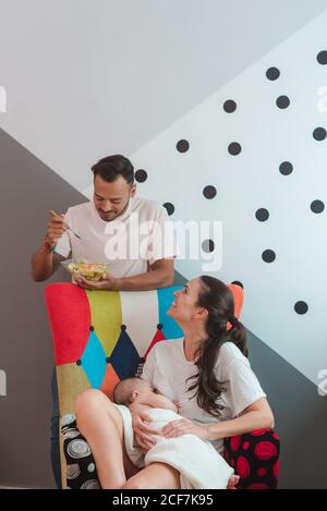 Black haired mother with sleepy baby ob chest with mobile phone in hand sitting on chair with father in casual wear eating salad Stock Photo