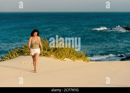 Tanned slim Woman in white shorts barefoot on sandy shore with plant on background of turquoise foamy waves in Fuerteventura, Las Palmas, Spain Stock Photo