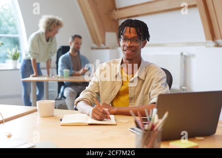 Portrait of African young man in eyeglasses sitting at his workplace smiling at camera while making notes in his note pad at office Stock Photo