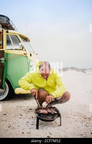 Tanned man cooking on camping stove beside parked mini van Stock Photo